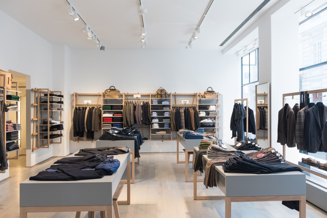 Modern retail clothing store interior with wooden display shelves, hanging garments, and minimalist white walls.