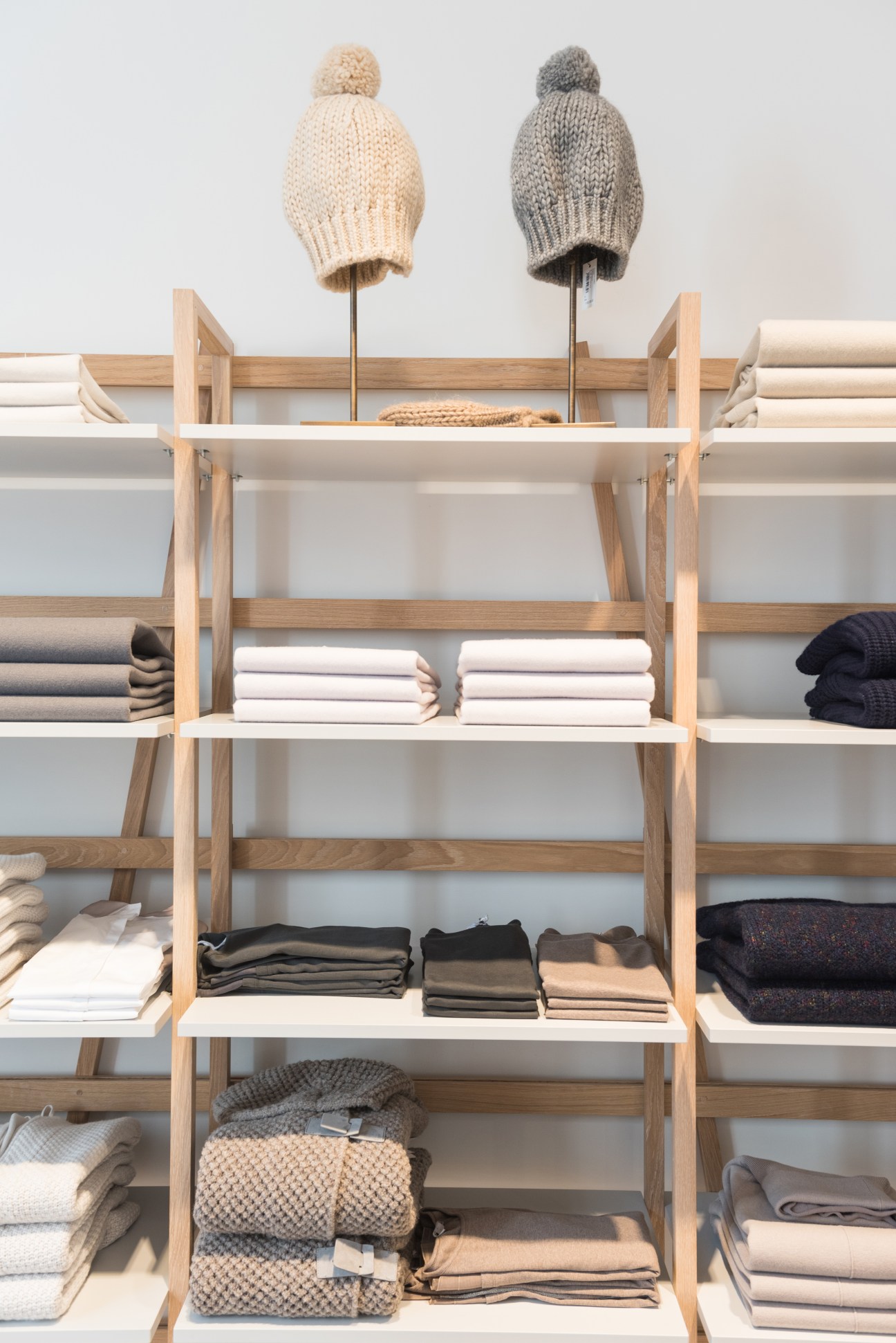 Light wood display shelving with folded knitwear and two knit beanies with pom-poms displayed on stands.