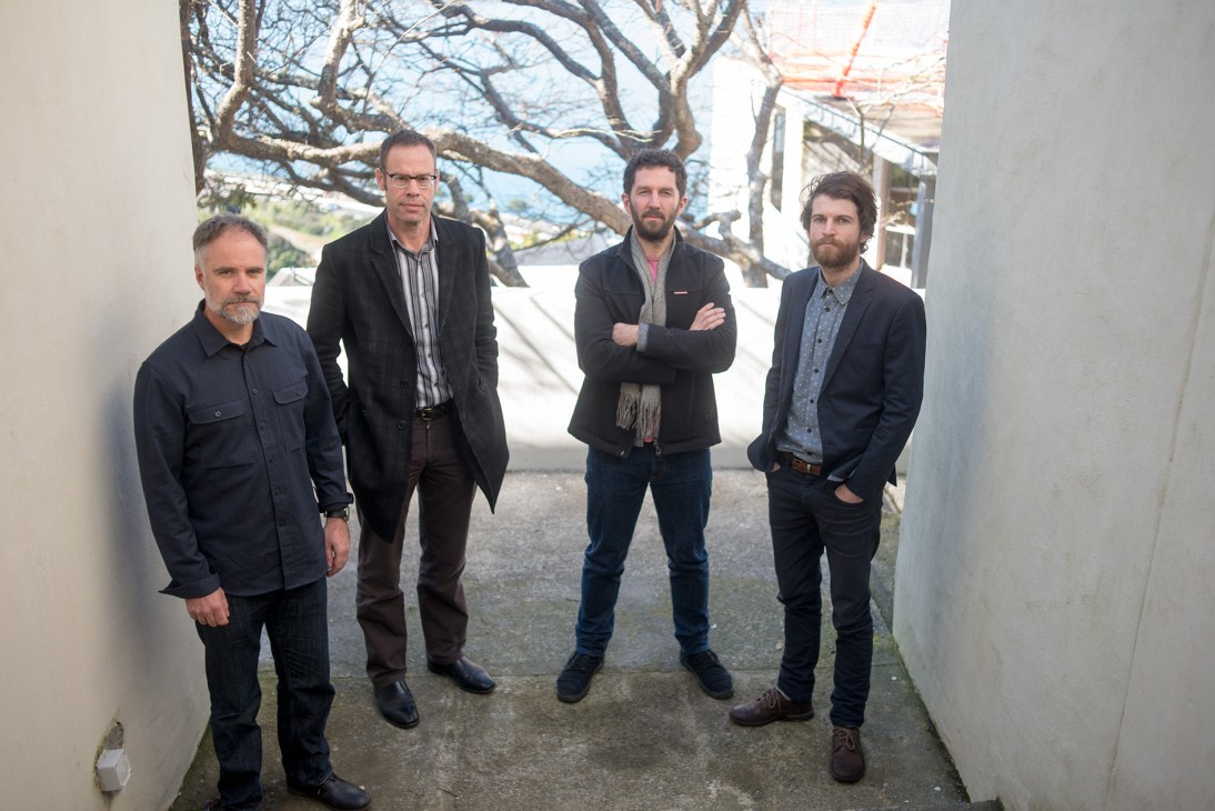 Four men standing in a white corridor with bare trees visible through a window behind them.