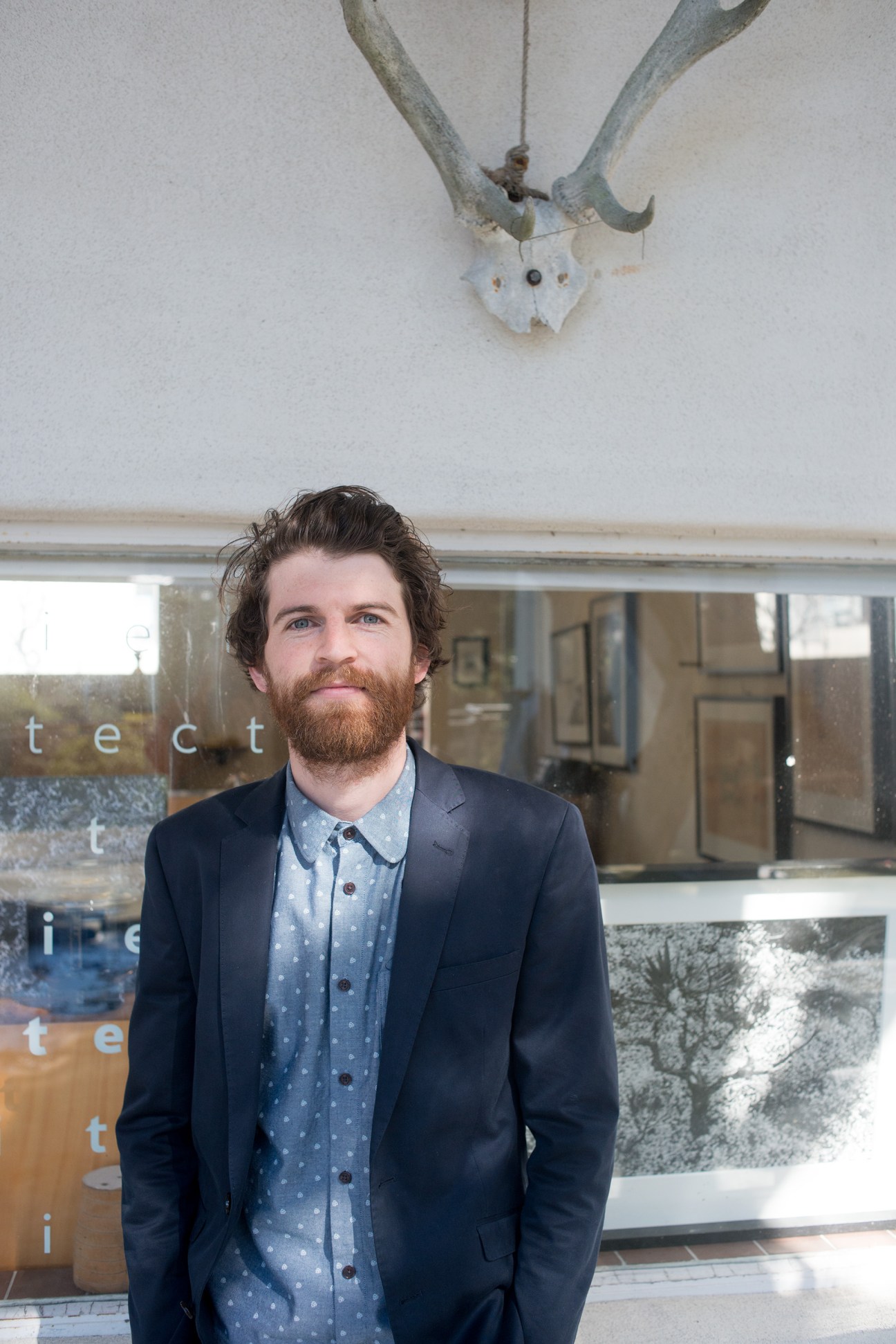 Man with beard wearing navy blazer and patterned shirt stands in front of window with animal skull mounted above.