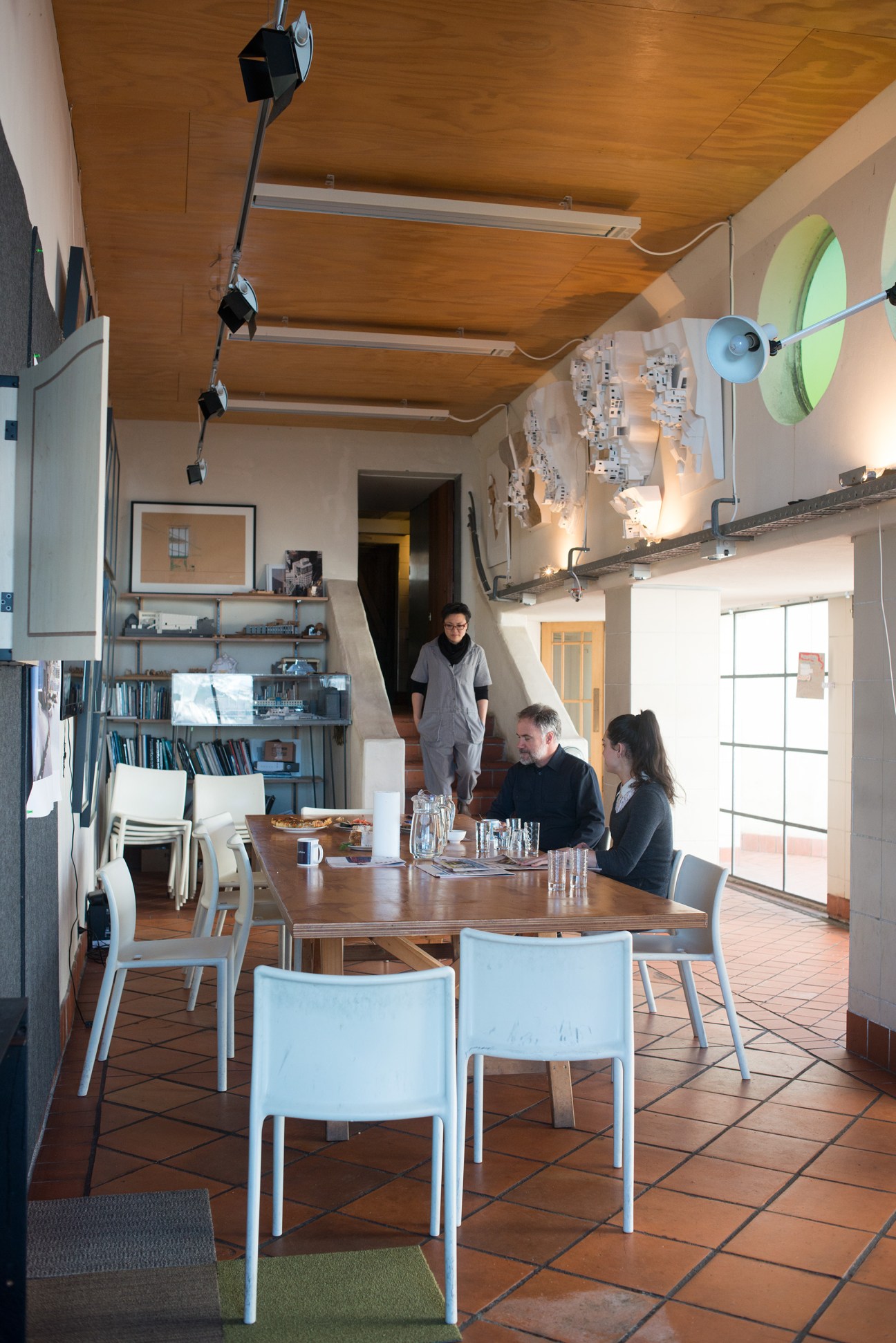 Interior of an architecture office with wooden ceiling, staff at a dining table, bookshelves, and design materials on walls.