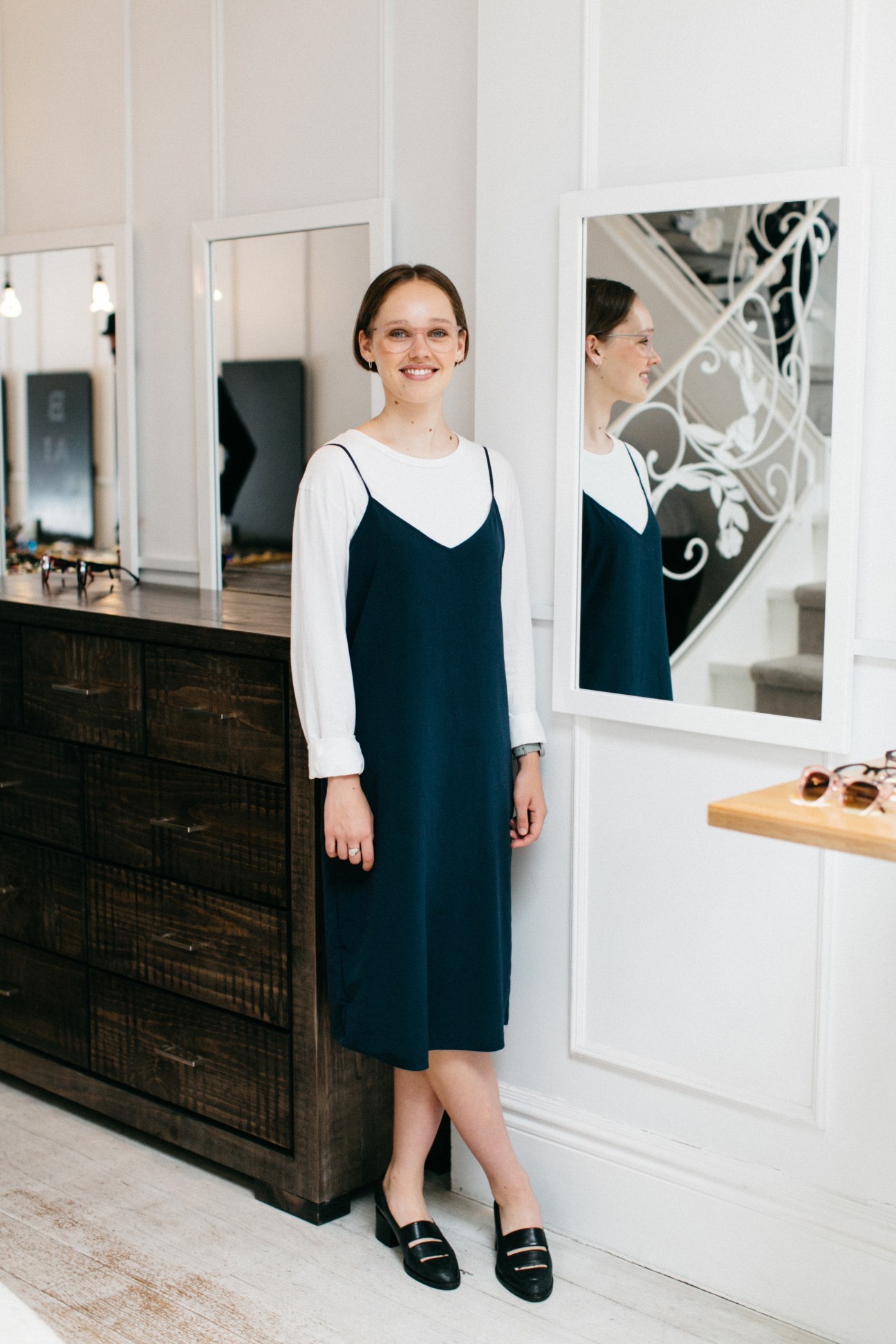 Young woman smiling in Bailey Nelson eyewear shop wearing navy slip dress and white long-sleeve shirt.