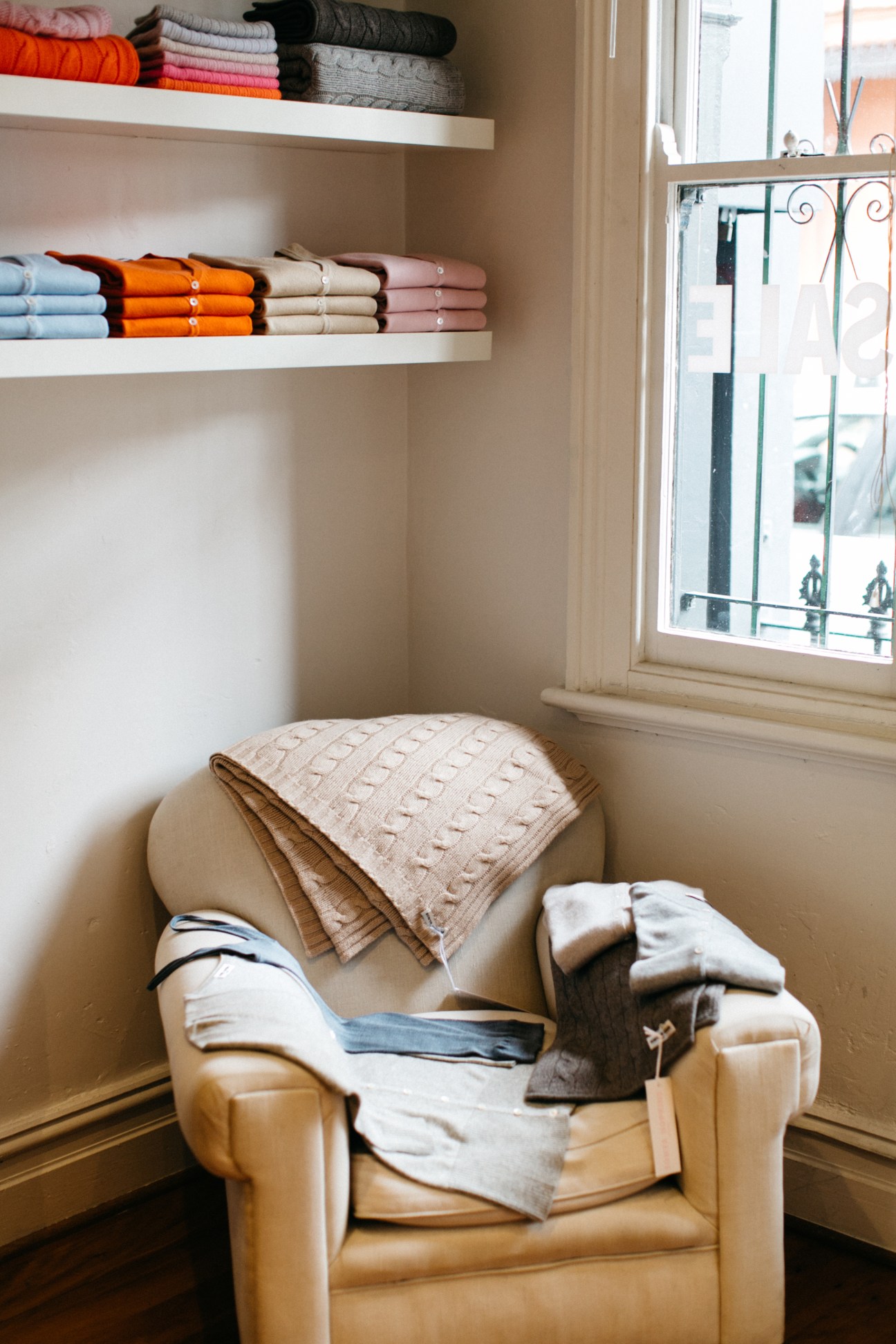 Colorful folded knitwear displayed on white shelves above a tan leather chair by a window.