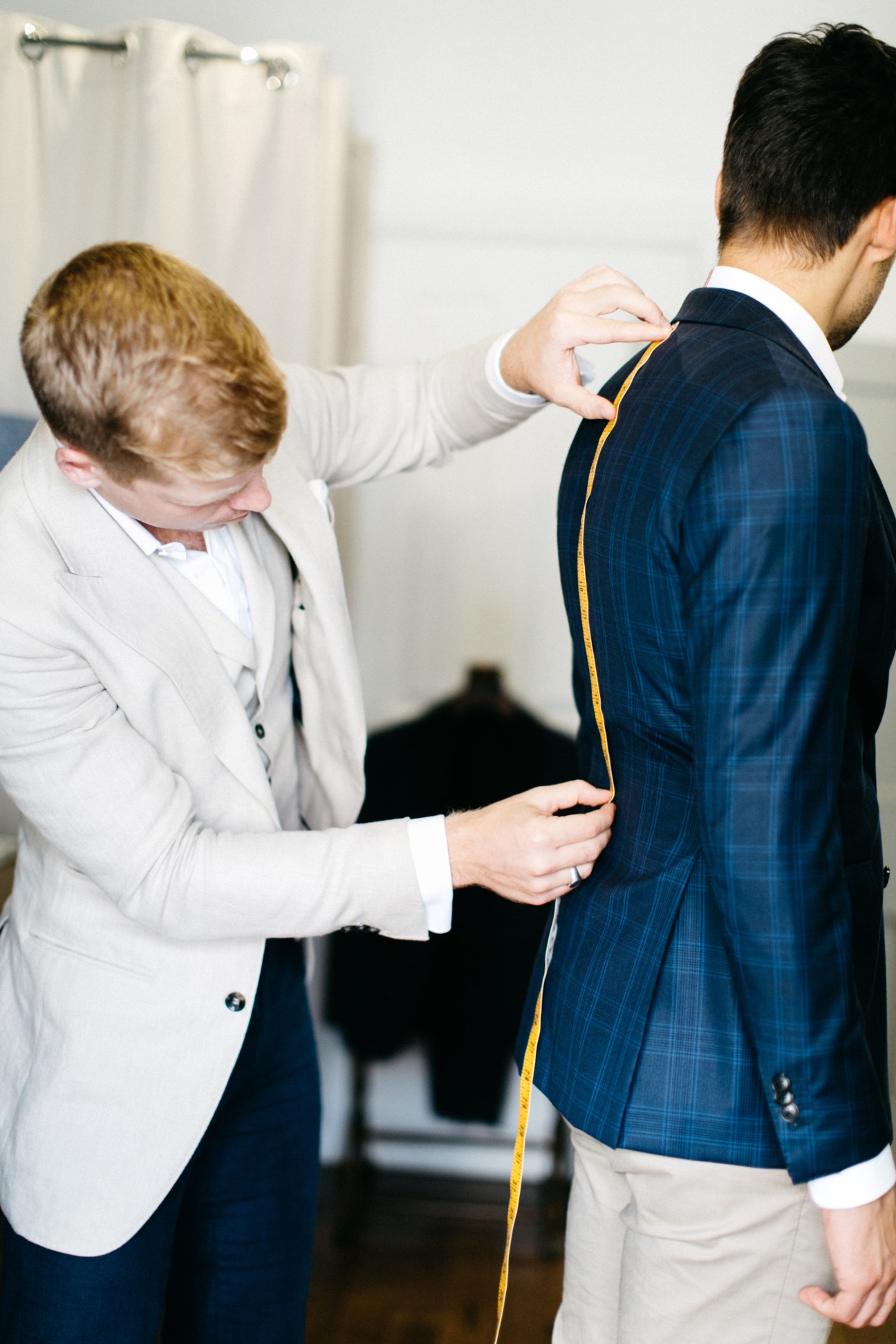 A tailor in a white blazer measures a customer wearing a blue pinstriped jacket at Belancé menswear shop.