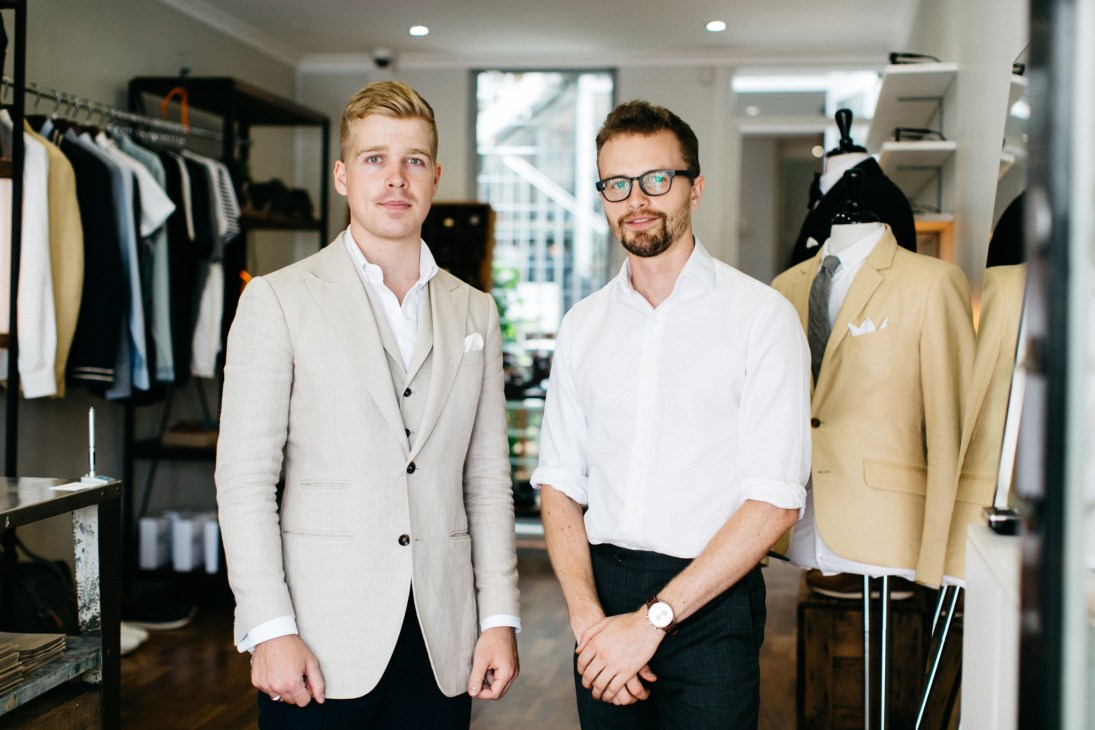 Two men stand in a clothing retail space with garments displayed on racks and a dress form behind them.