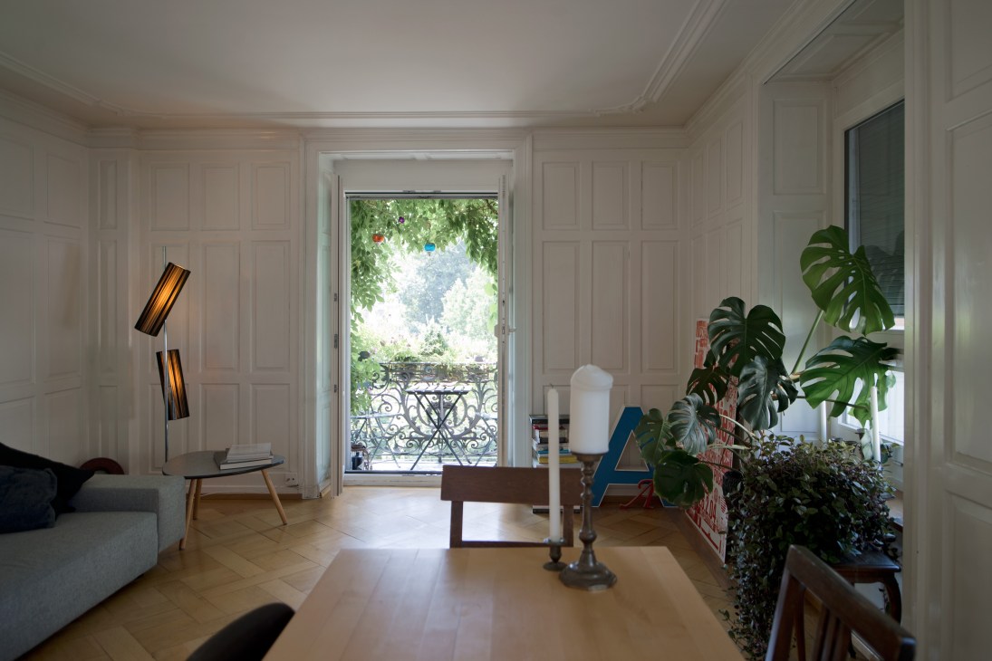 Minimalist apartment interior with white paneled walls, wooden floors, potted plants, and modern furnishings opening onto a leafy balcony.