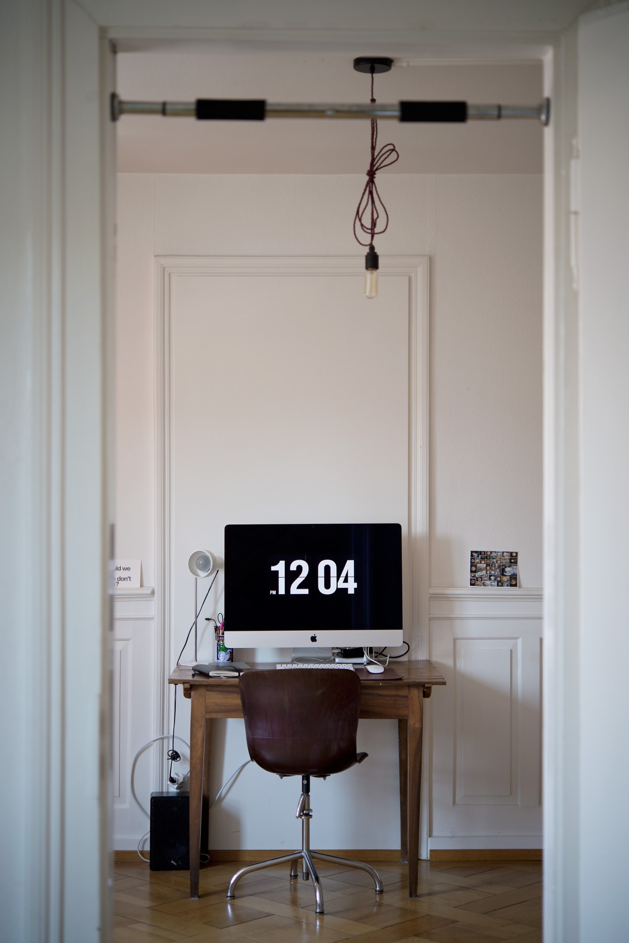 Minimalist home office with wooden desk, iMac displaying 12:04, leather chair, and pendant lamp above.