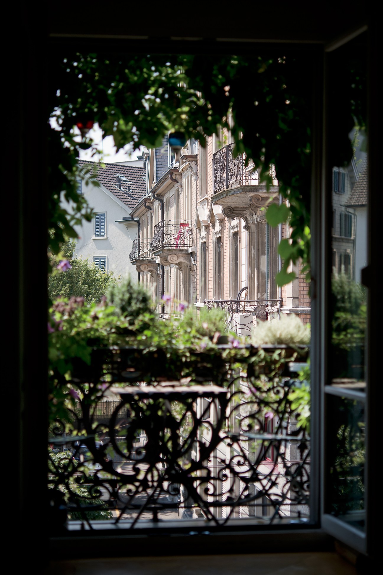 View through a balcony door framed by green foliage onto a Zurich street with ornate historic buildings with wrought-iron railings.