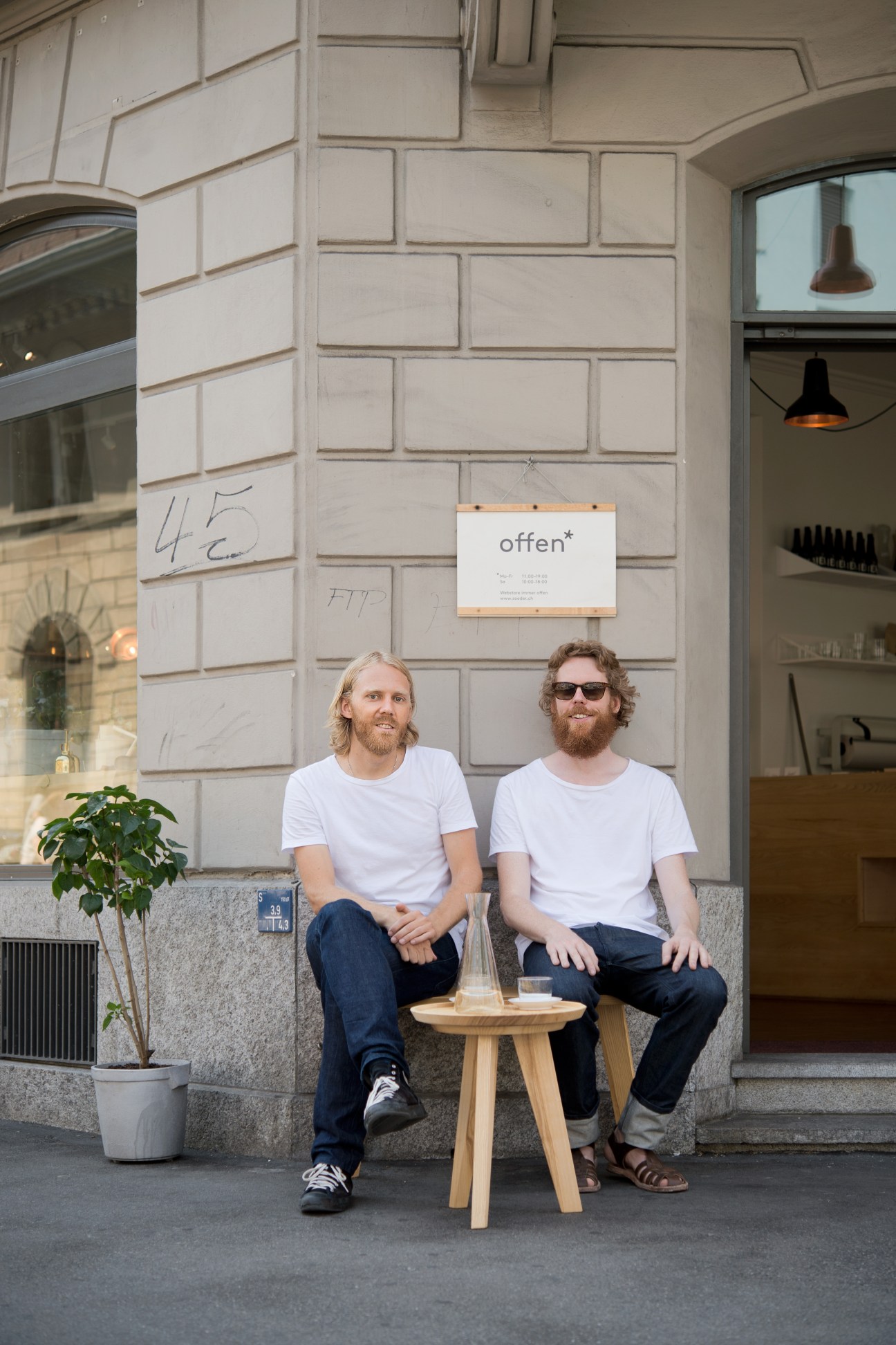 Two men in white t-shirts and dark jeans sit on a bench outside a storefront with a sign reading 'offer*'