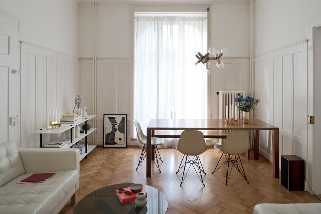 Minimalist apartment interior with wooden dining table, beige chairs, and white panelled walls with pendant lighting.