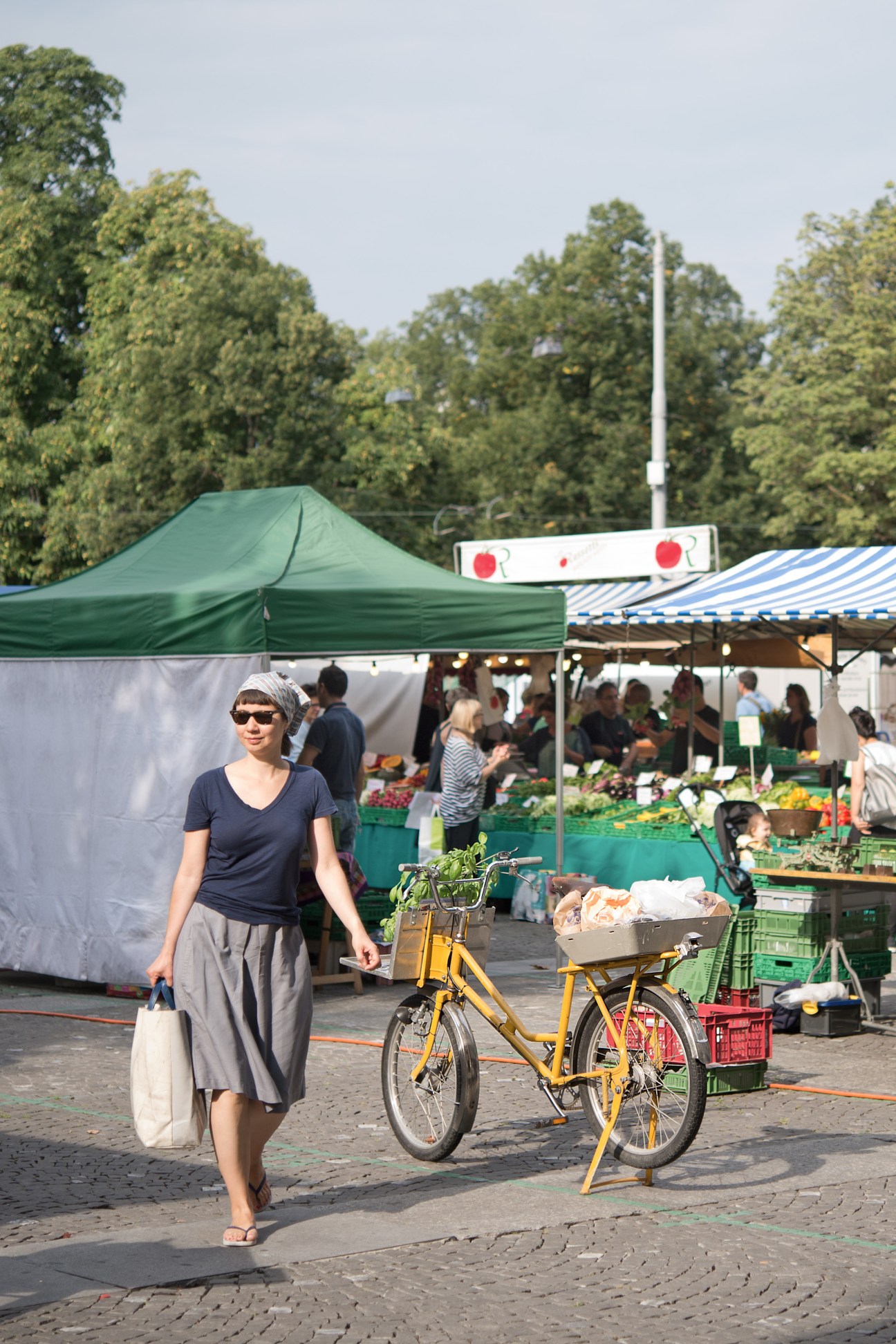 Woman with yellow bicycle at Helvetiaplatz market with vendor tents and trees in background.