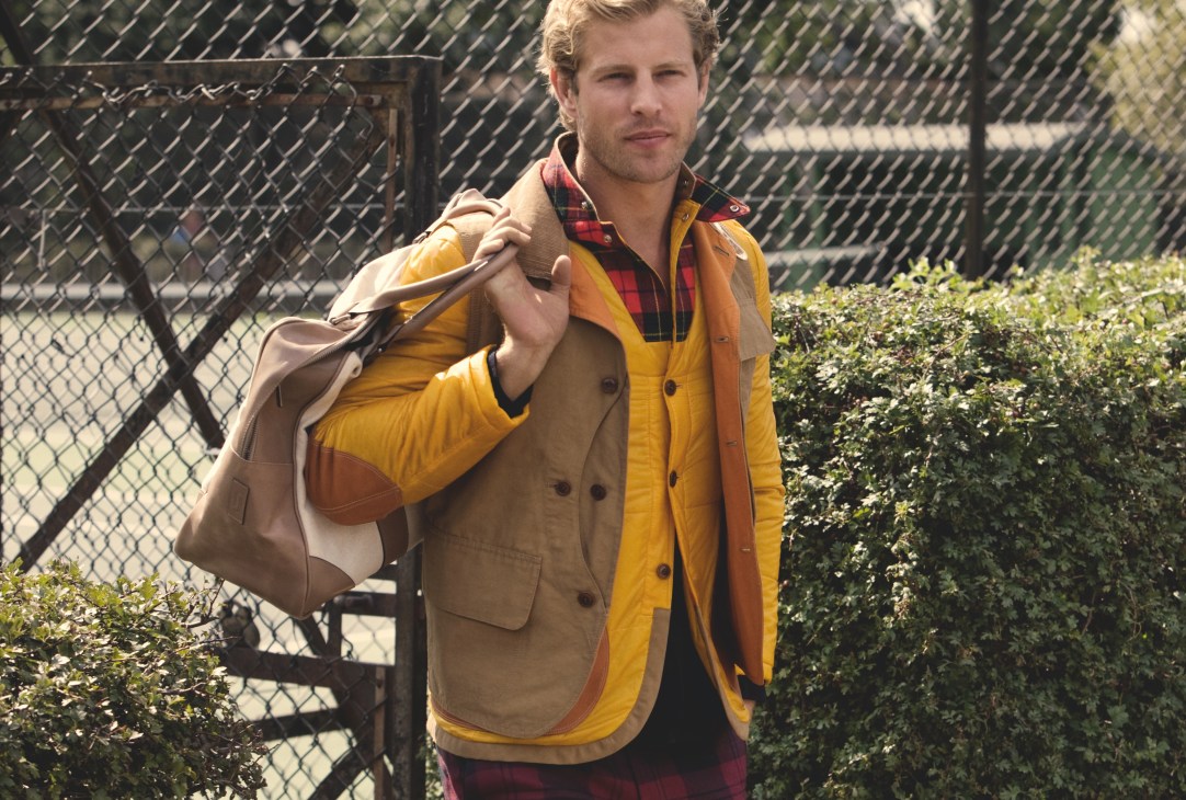 Man in yellow and tan jacket with plaid shirt holds brown leather bag at tennis court.