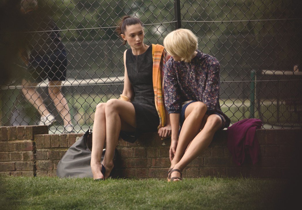 Two women sitting on a brick bench by a tennis court fence wearing stylish off-court outfits.
