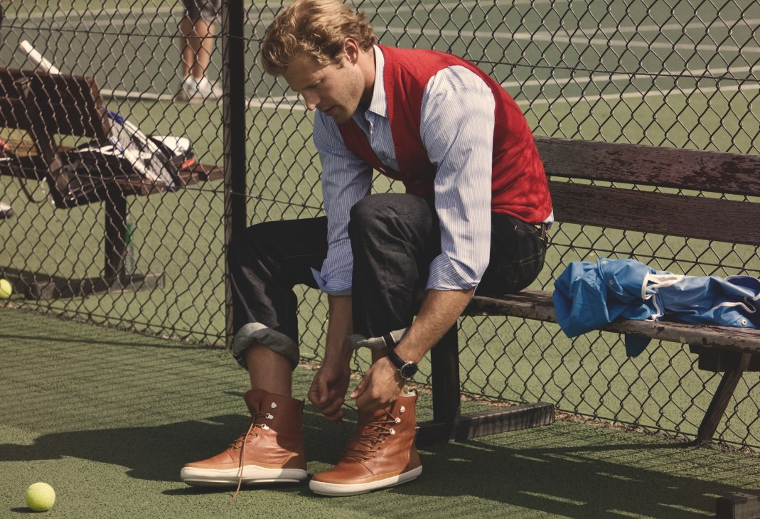 Man sitting on bench at tennis court wearing red vest, blue shirt, and tan leather boots.