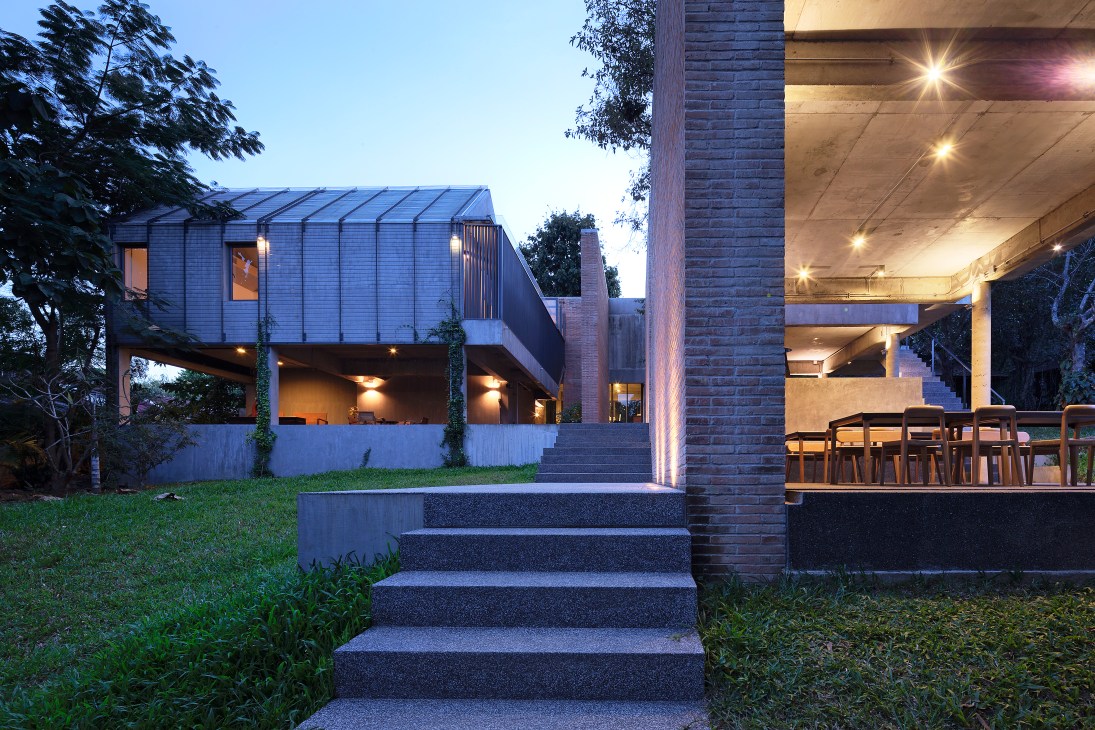 Modern residential house elevated on stilts with blue metal cladding and brick elements, lit at dusk with lawn and trees.