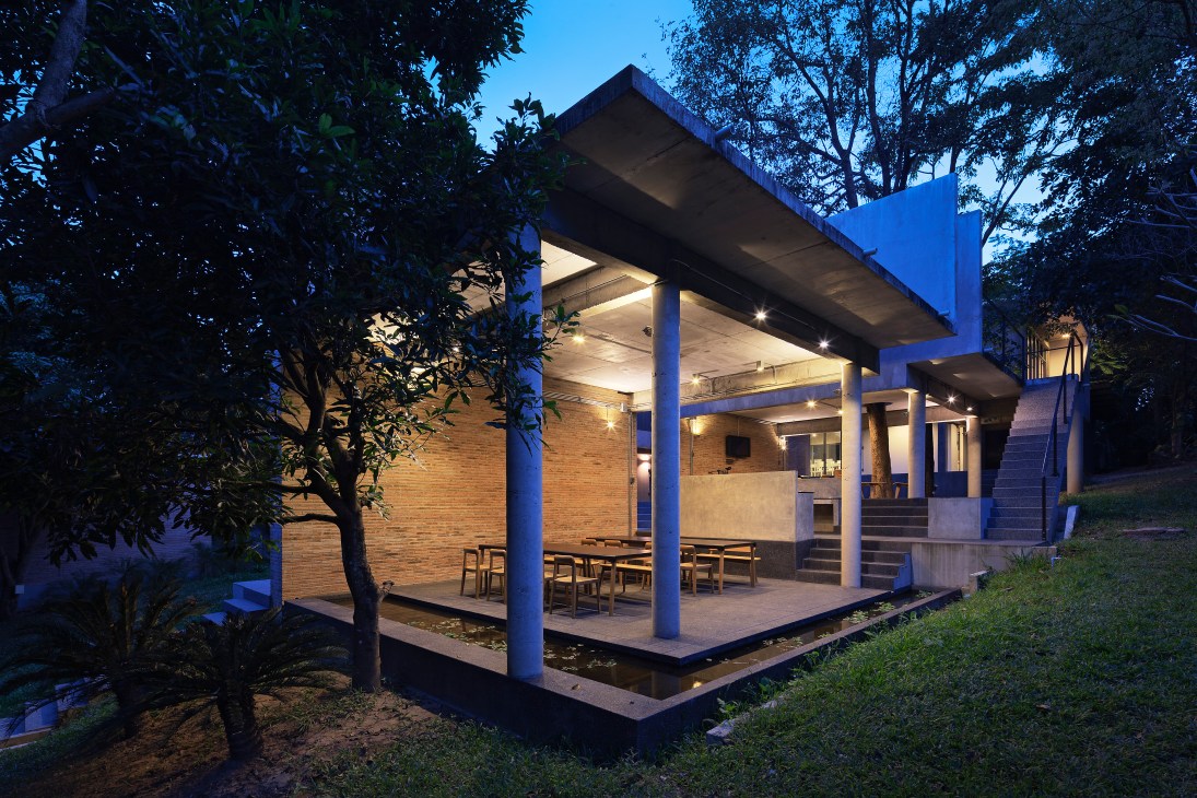 Modern raised house with blue pillars and brick walls illuminated at dusk, surrounded by trees.