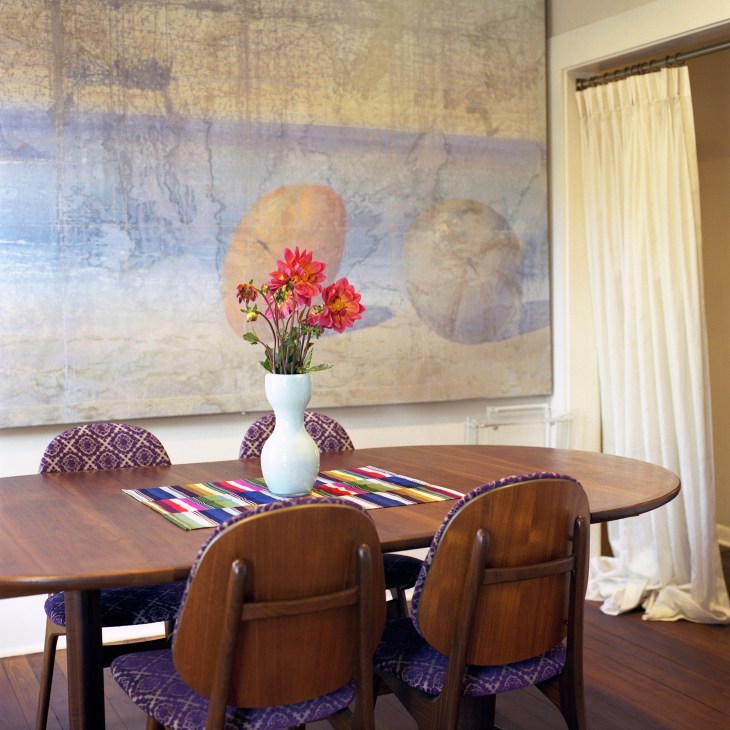 A minimalist dining room with a wooden oval table, mid-century chairs, and a white vase of pink flowers against textured walls.