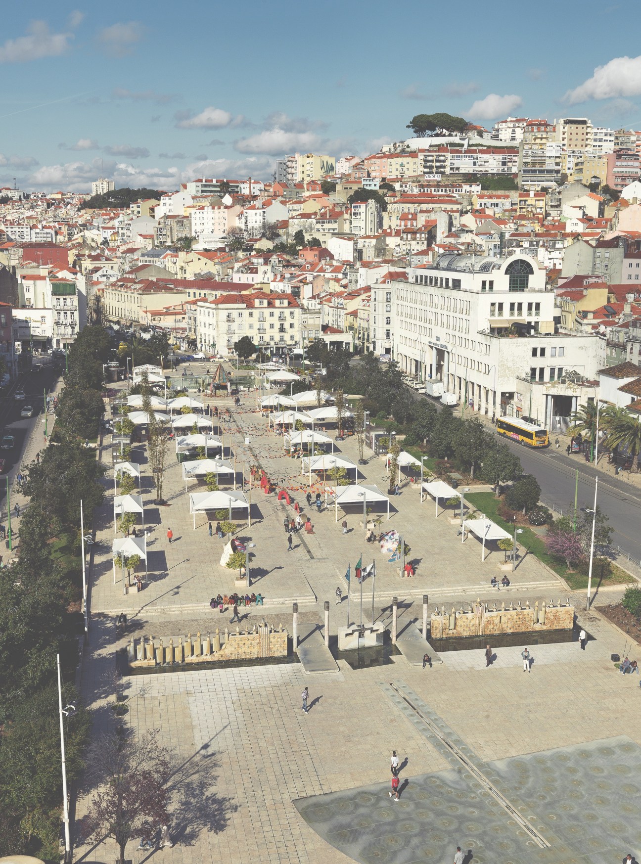Aerial view of Largo do Intendente plaza in Lisbon's Mouraria neighbourhood with white market tents, surrounding historic buildings and hills.