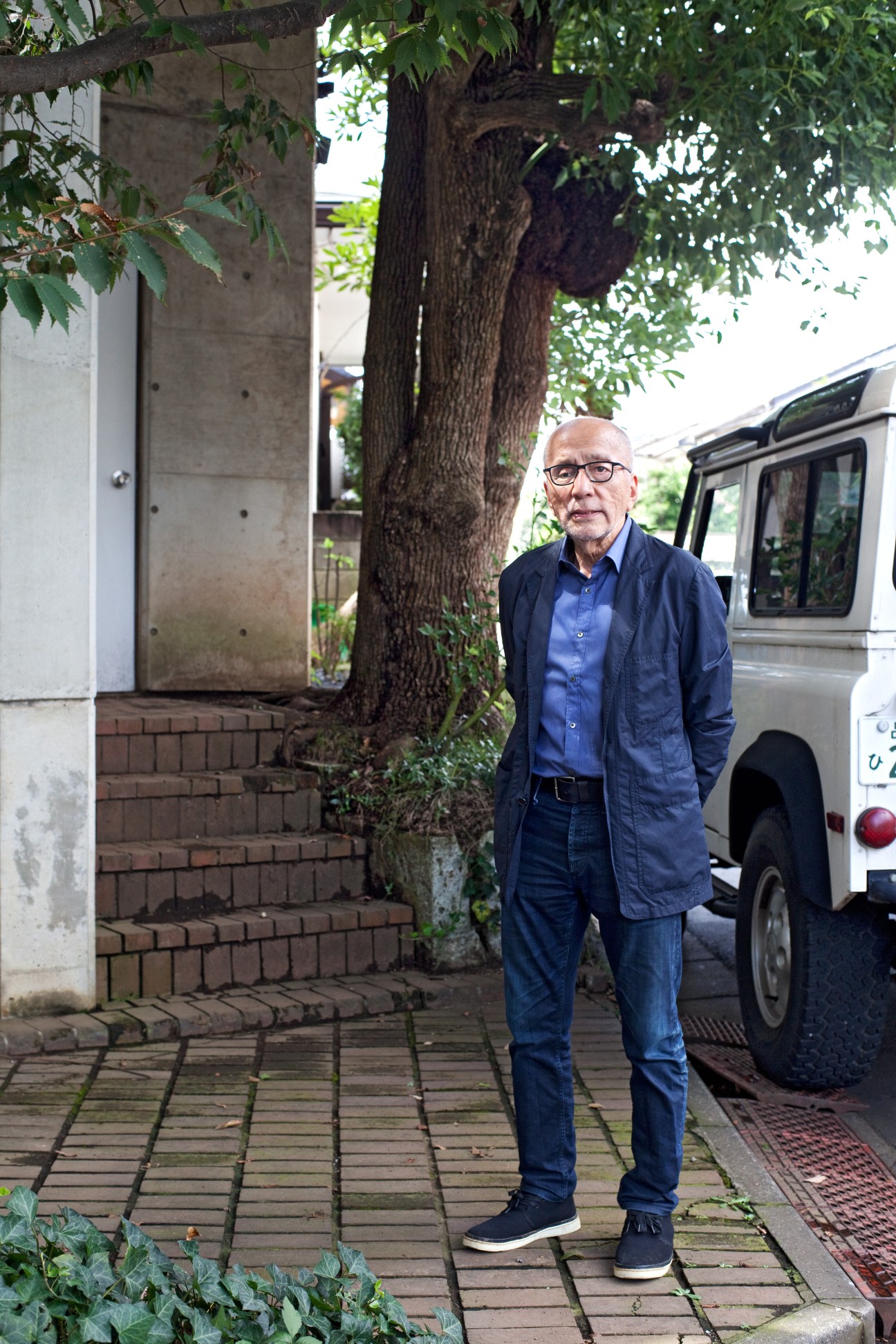 Tsutomu Abe standing outdoors in blue clothing next to his white Land Rover with a large tree in the background