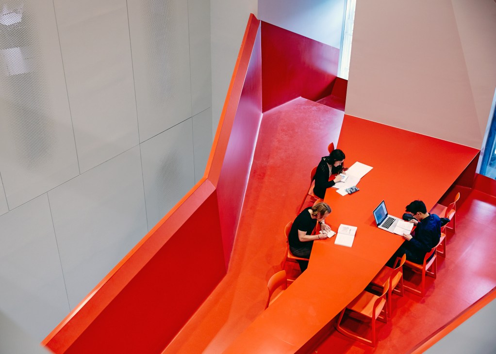 Students work at tables in the Lovell Chen lounge, a brightly colored red and orange interior space at the Melbourne School of Design.