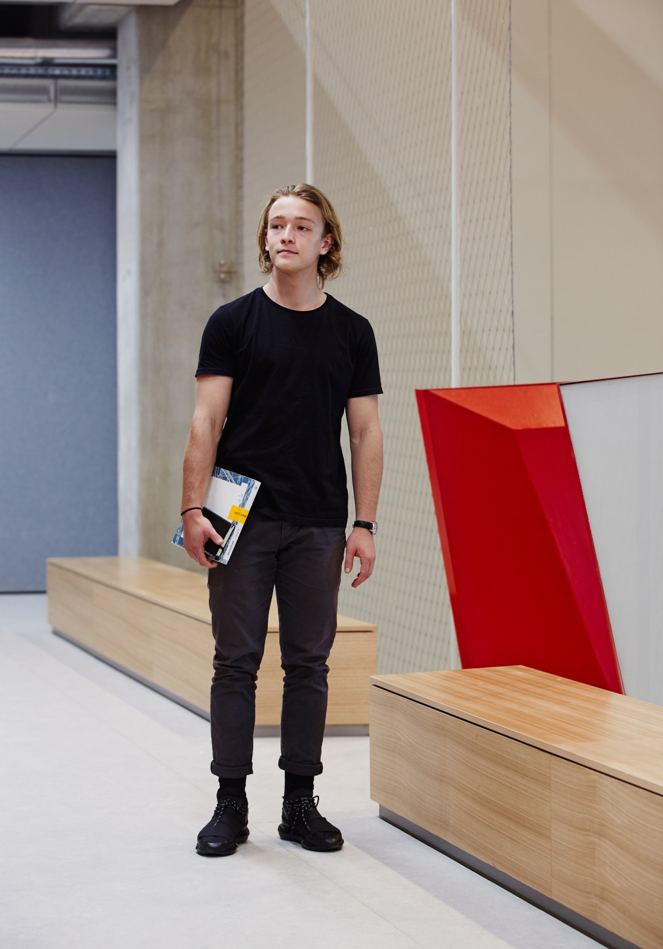 Student Sam Chesbrough standing in the Melbourne School of Design atrium with wooden bench and red wall element visible.