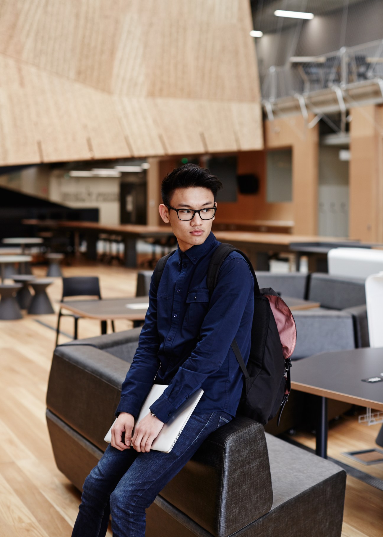 Student Lawson Heah sits in the Melbourne School of Design atrium holding a laptop, wearing glasses and a navy jacket.