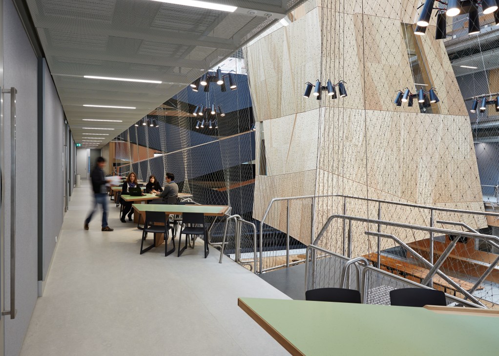 Students work at tables on a second-level balcony overlooking the Melbourne School of Design atrium, with steel netting railings and suspended timber studios visible above.
