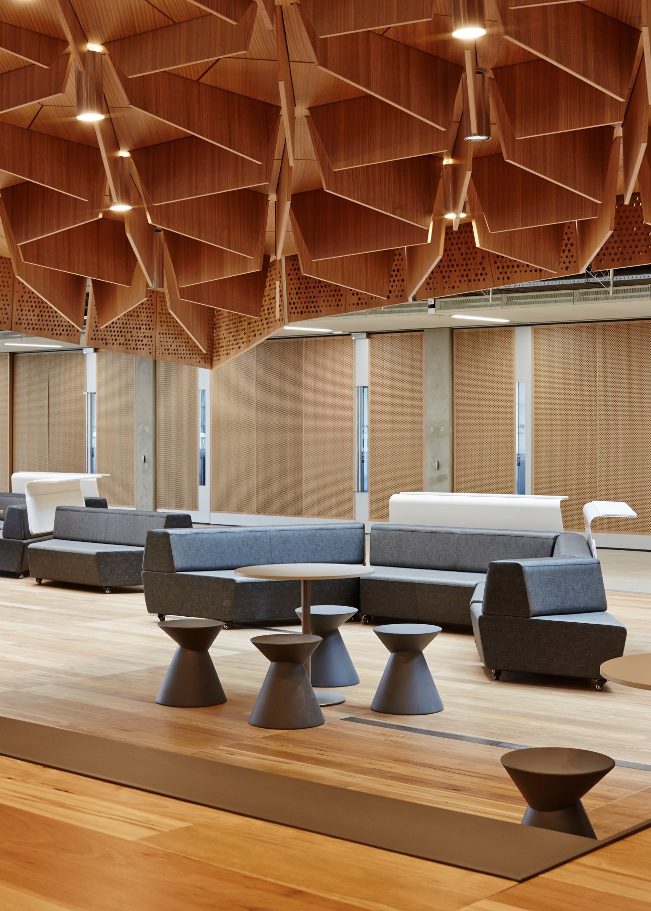 Modern lounge with grey upholstered seating, cone-shaped side tables, and timber coffered ceiling with pendant lights.