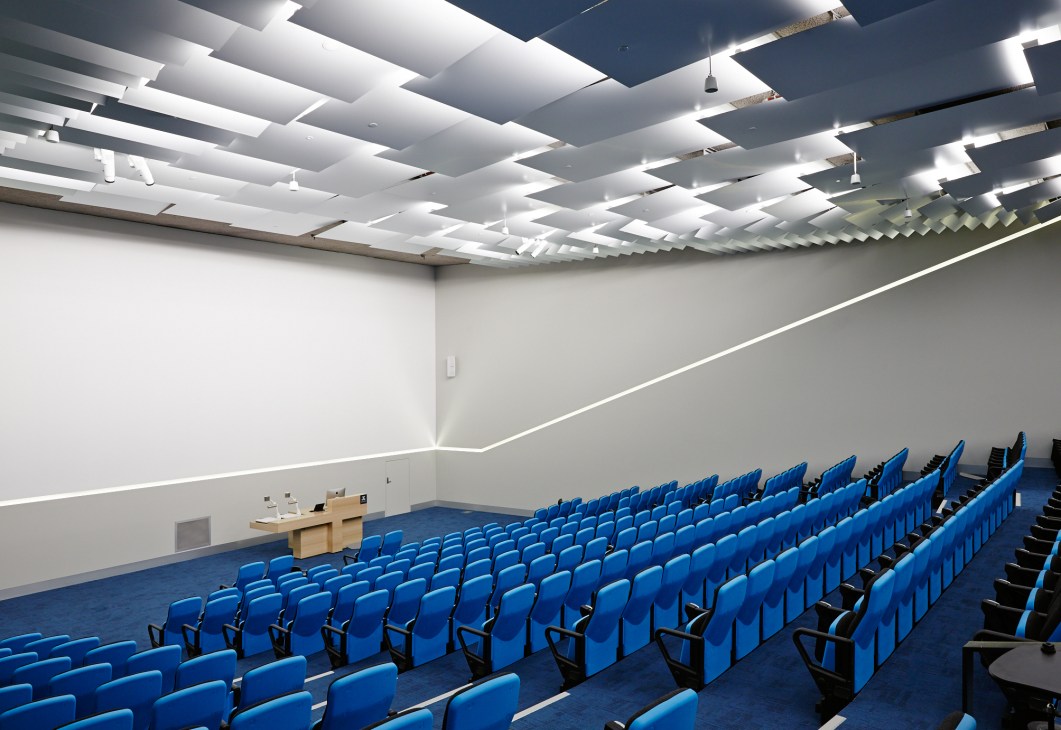 Empty lecture theatre with rows of blue seats facing a wooden podium and geometric white ceiling.