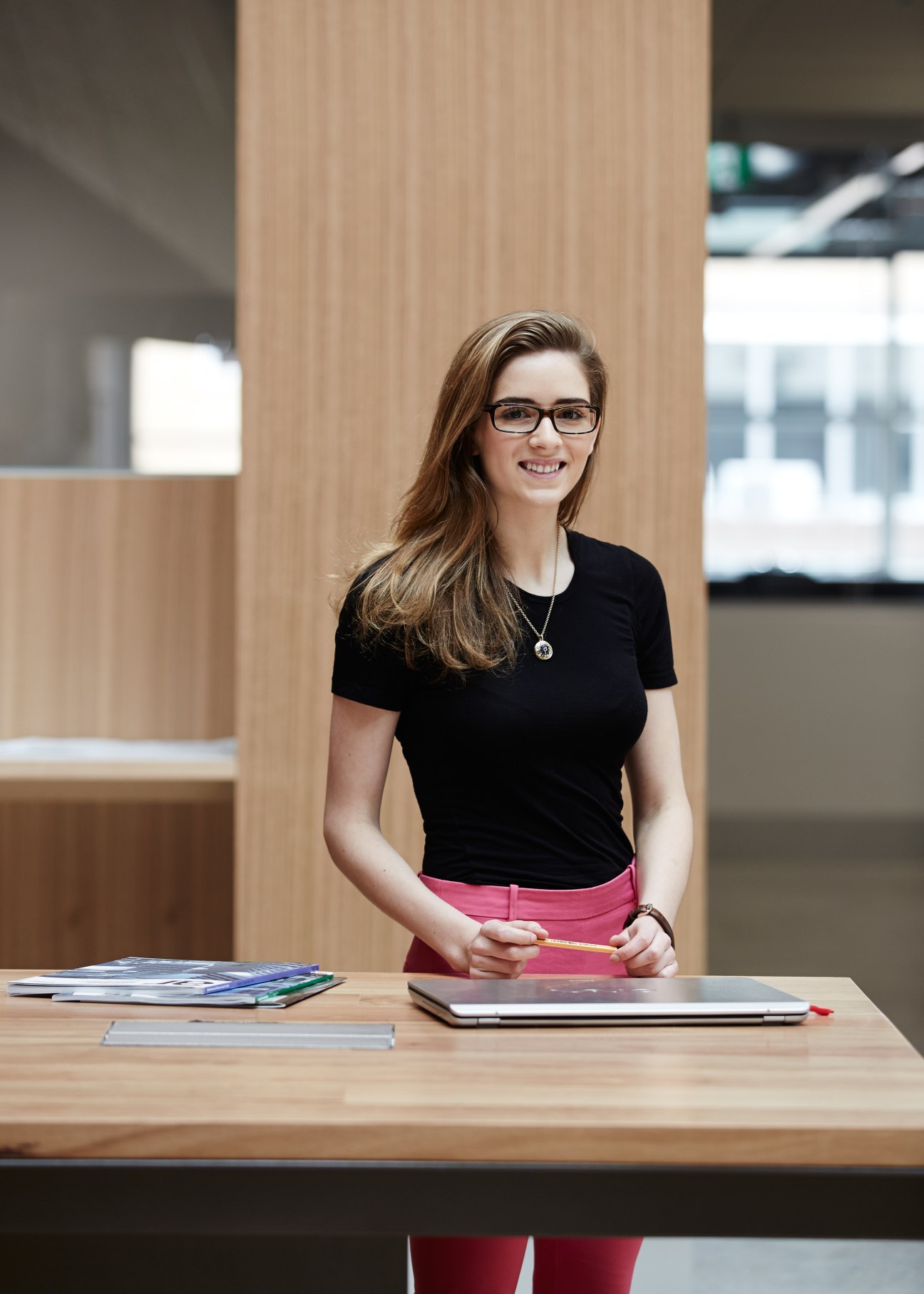 Student Olivia Gude stands at a wooden desk in the Melbourne School of Design atrium with timber architectural elements visible behind her.