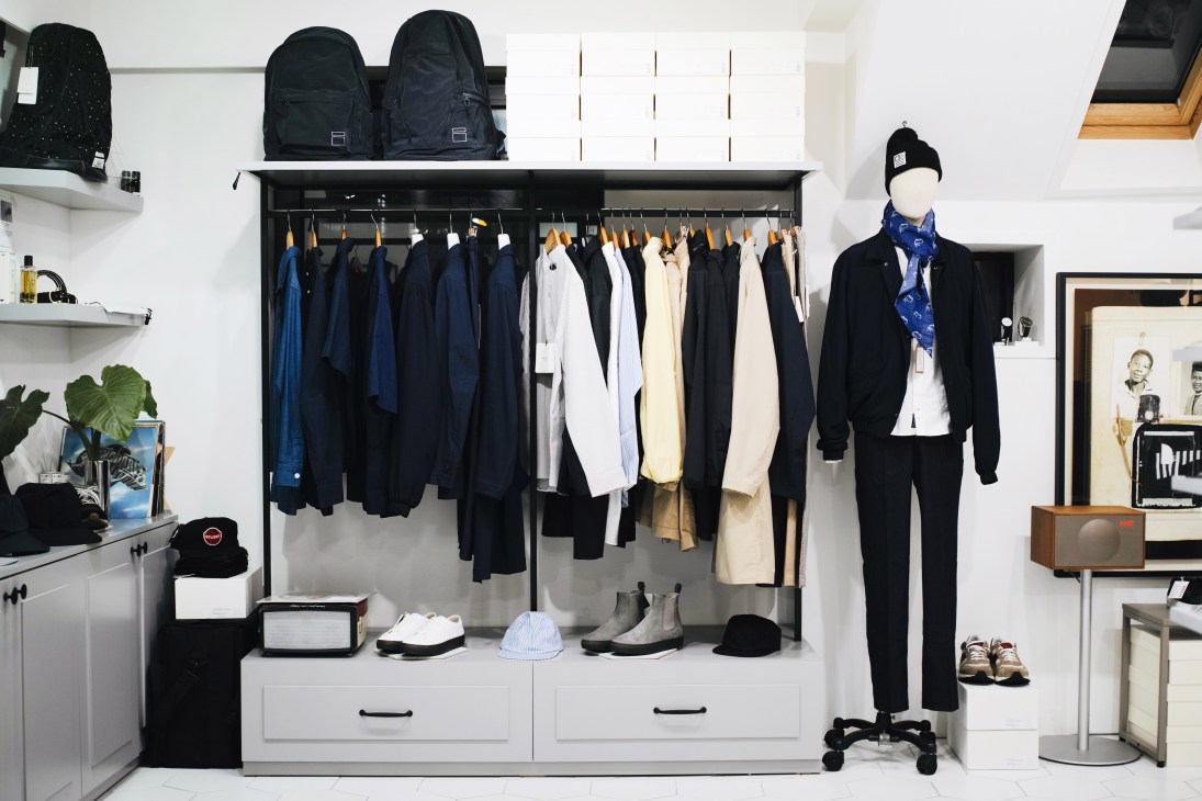 Clean menswear shop interior with clothing rails displaying navy, black and cream garments, a mannequin in tailored outfit, and displayed accessories on white shelving.