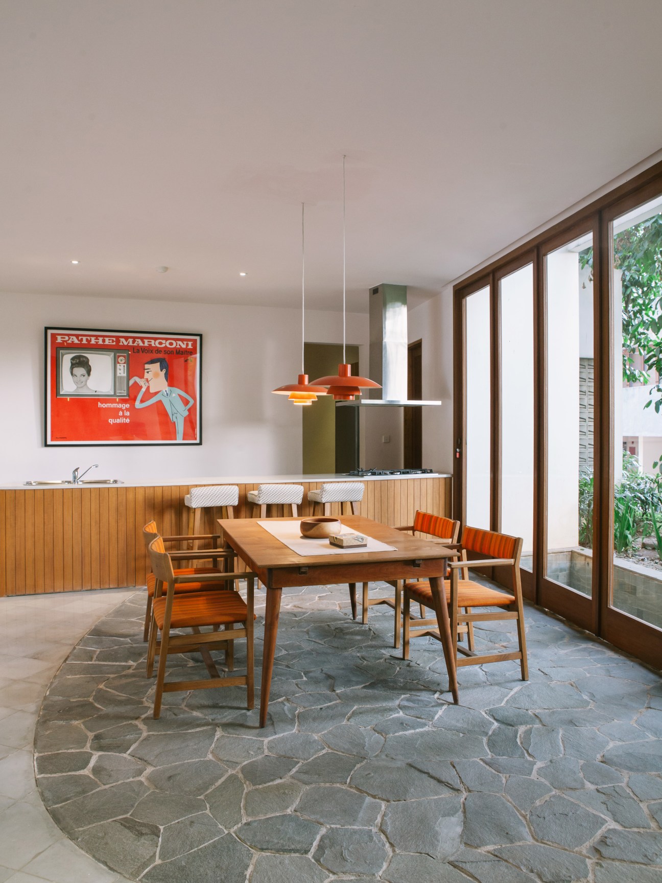 Modern dining room with wooden table, orange chairs, wood-paneled bar and vintage Pathe Marconi poster.