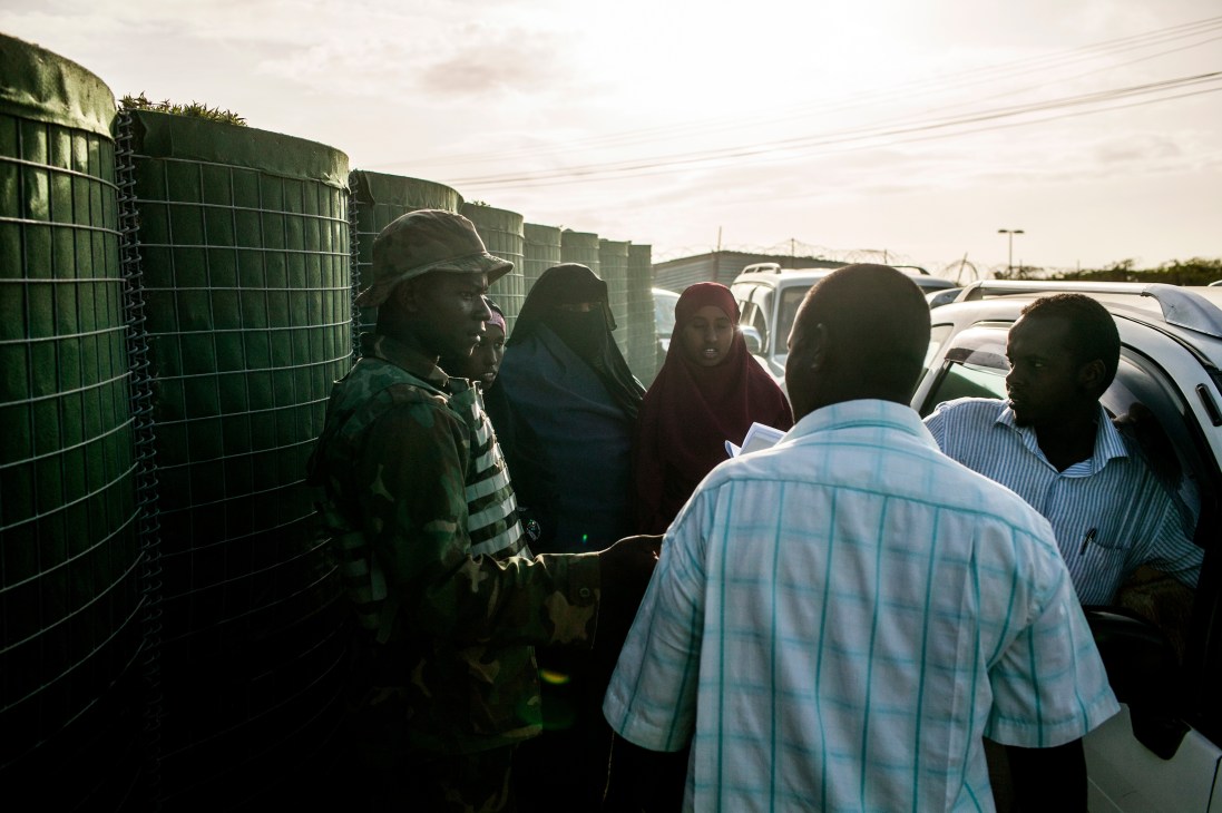 People pass through an Amisom security checkpoint at Mogadishu airport with green metal barriers and parked vehicles visible.