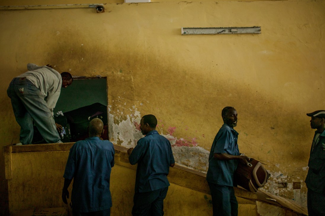 Baggage handlers in blue uniforms unload luggage in a deteriorated yellow-walled terminal room.