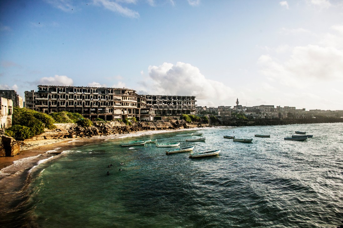 Mogadishu's war-damaged waterfront buildings overlook a beach with fishing boats anchored in turquoise waters.