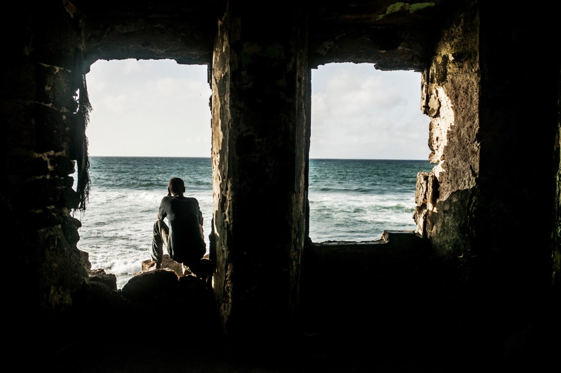 Person sitting in dark concrete ruins looking out toward the sea through two window openings.