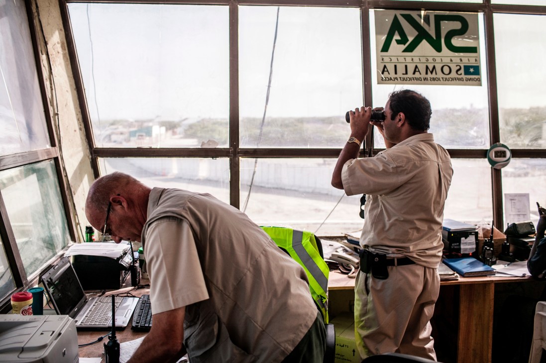 Two SKA employees monitor airport operations from a watchtower overlooking Mogadishu's landscape through large windows.