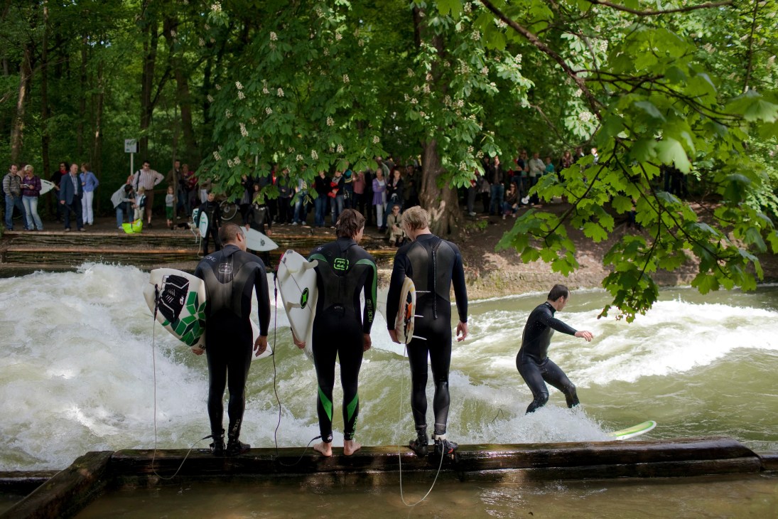 Surfers in wetsuits prepare to enter artificial wave at English Garden park in Munich.