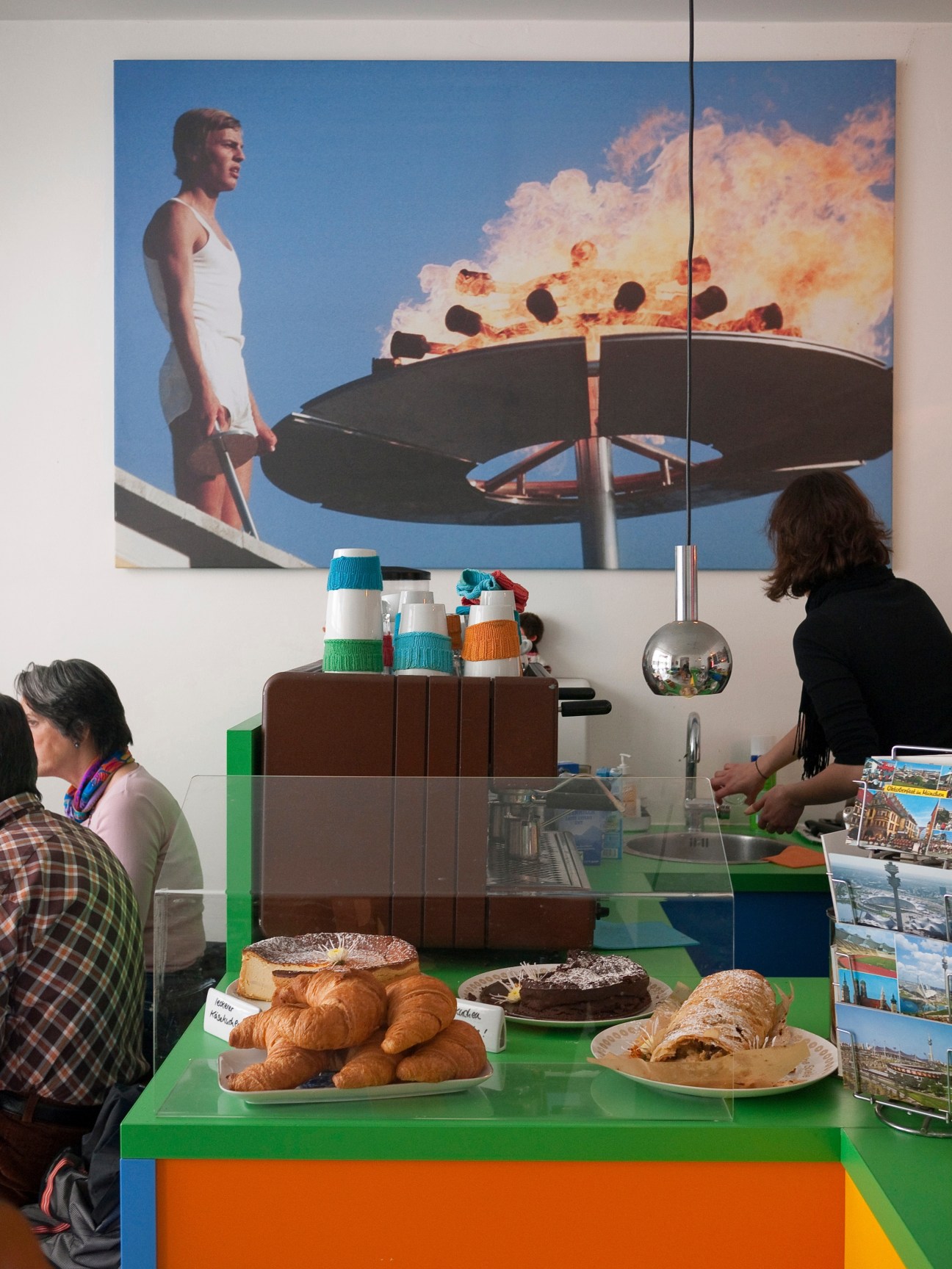 Interior of München 72 café with customers at colorful counter displaying pastries and cakes beneath vintage 1972 Olympics artwork.