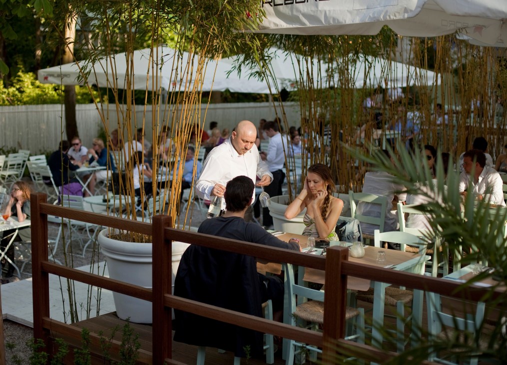 Outdoor waterside restaurant with server attending to diners under bamboo screening and white umbrellas.