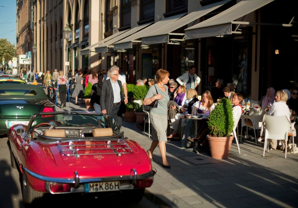 Pedestrians walk past outdoor café seating on a Munich street with historic architecture and awnings.