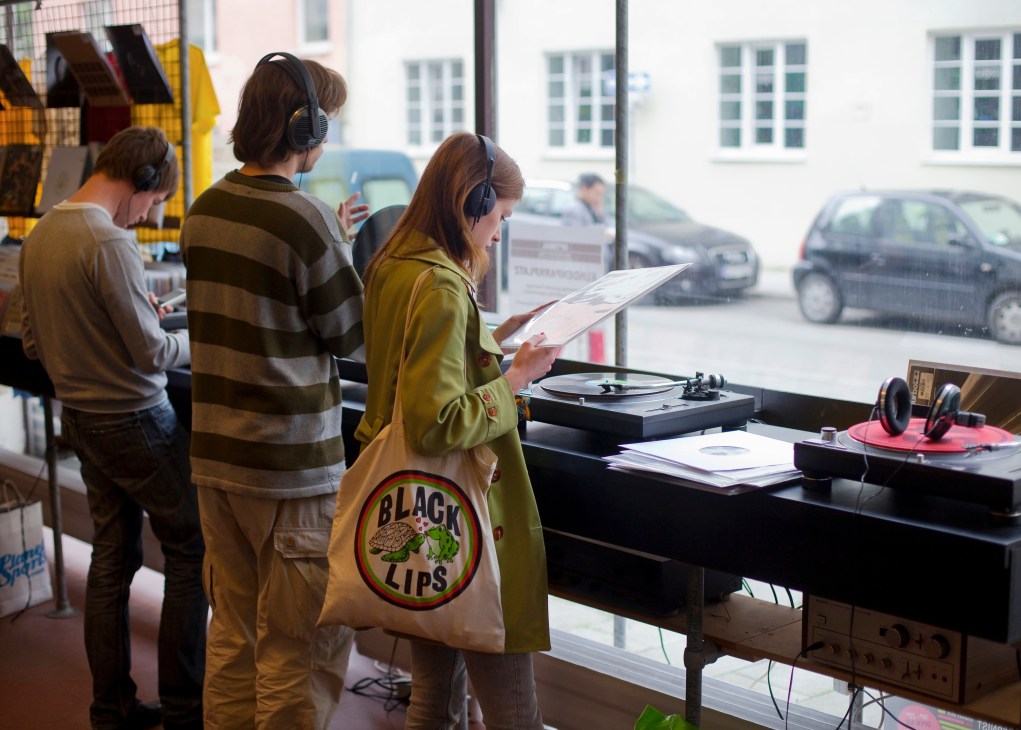 People wearing headphones browse vinyl records at Echt Optimal Schallplatten record store with turntable and equipment visible on counter.