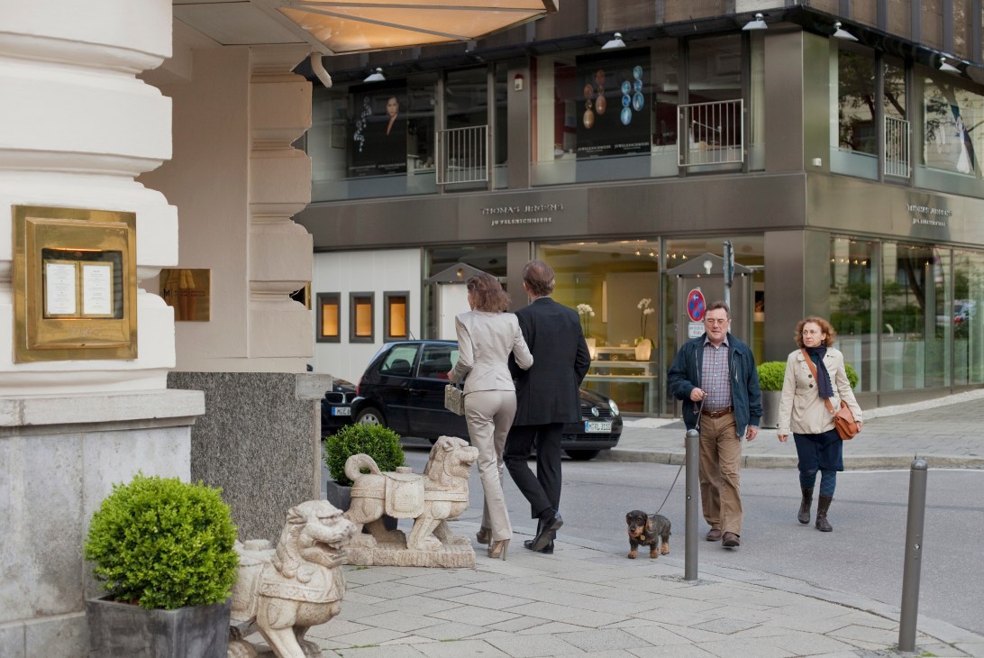 Pedestrians and a small dog on a Munich street outside the Mandarin Oriental hotel with storefronts and decorative planters.