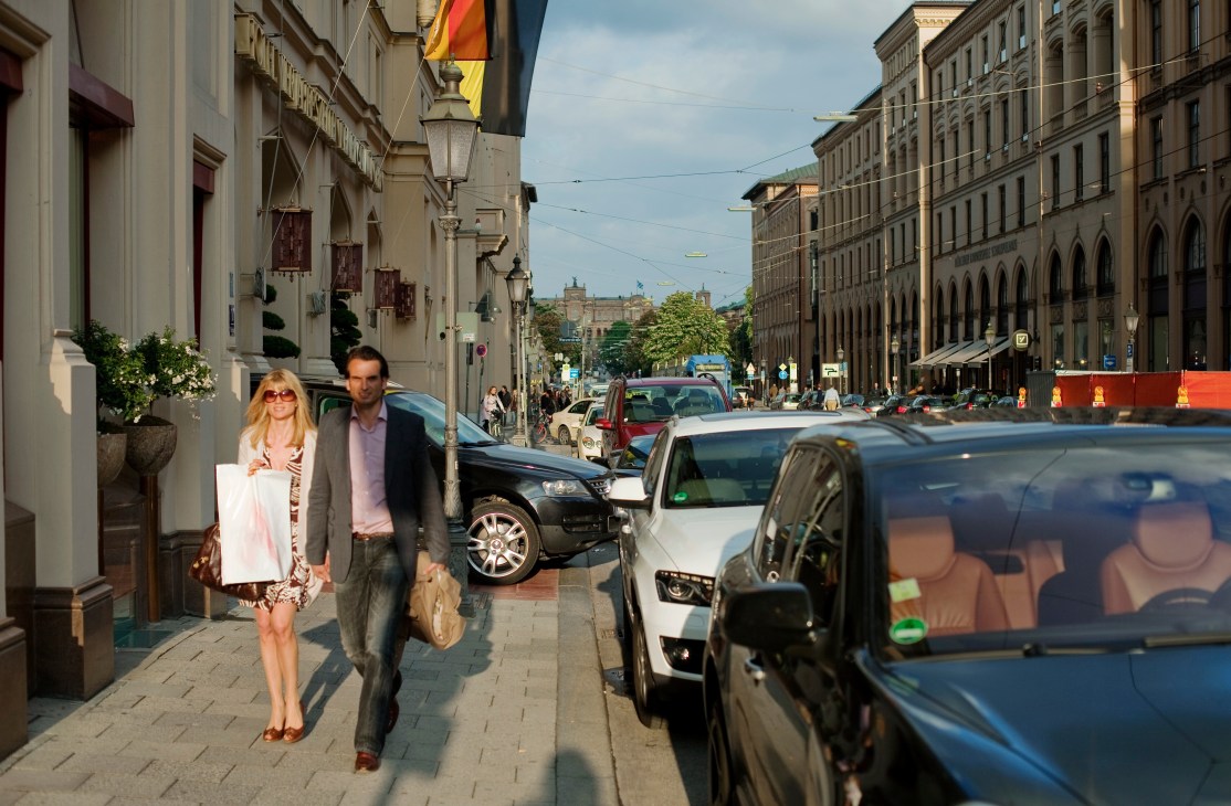 Two pedestrians walk on Maximilianstrasse in Munich past parked cars and historic buildings.