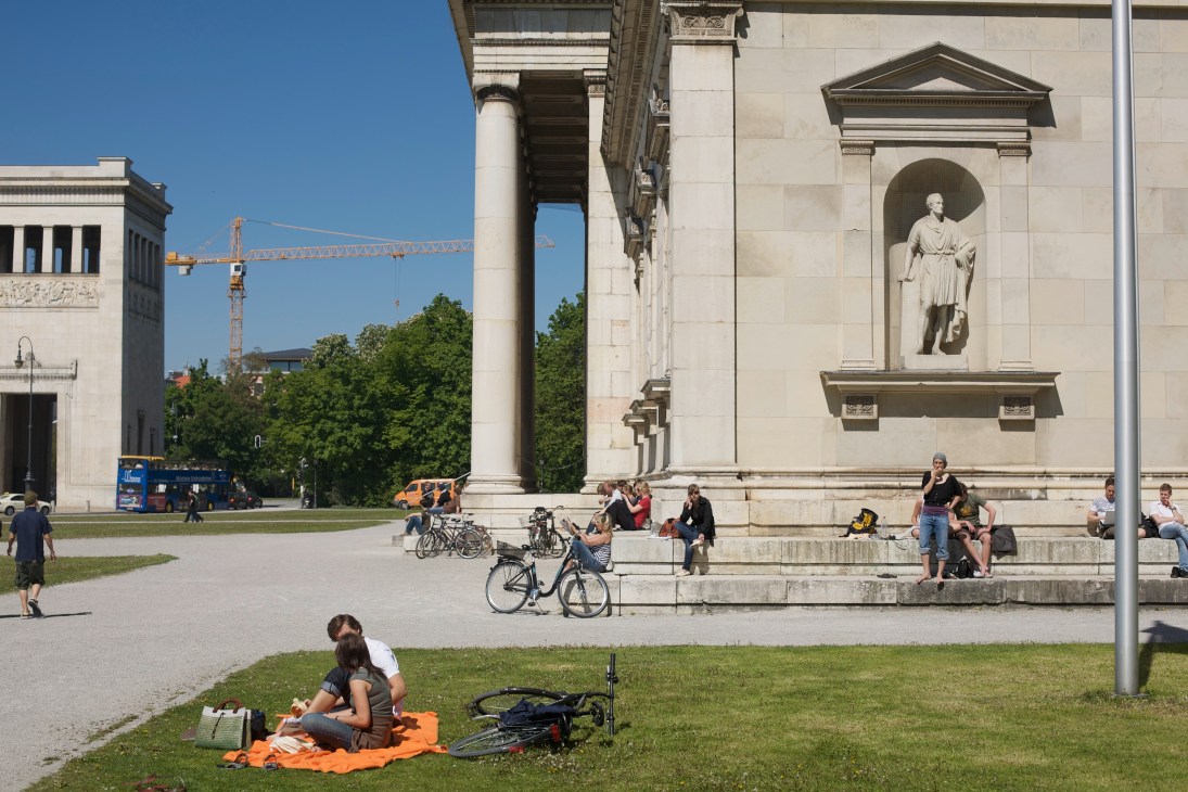 Classical museum courtyard with columned building, statue niche, and people relaxing on grass with bicycles.