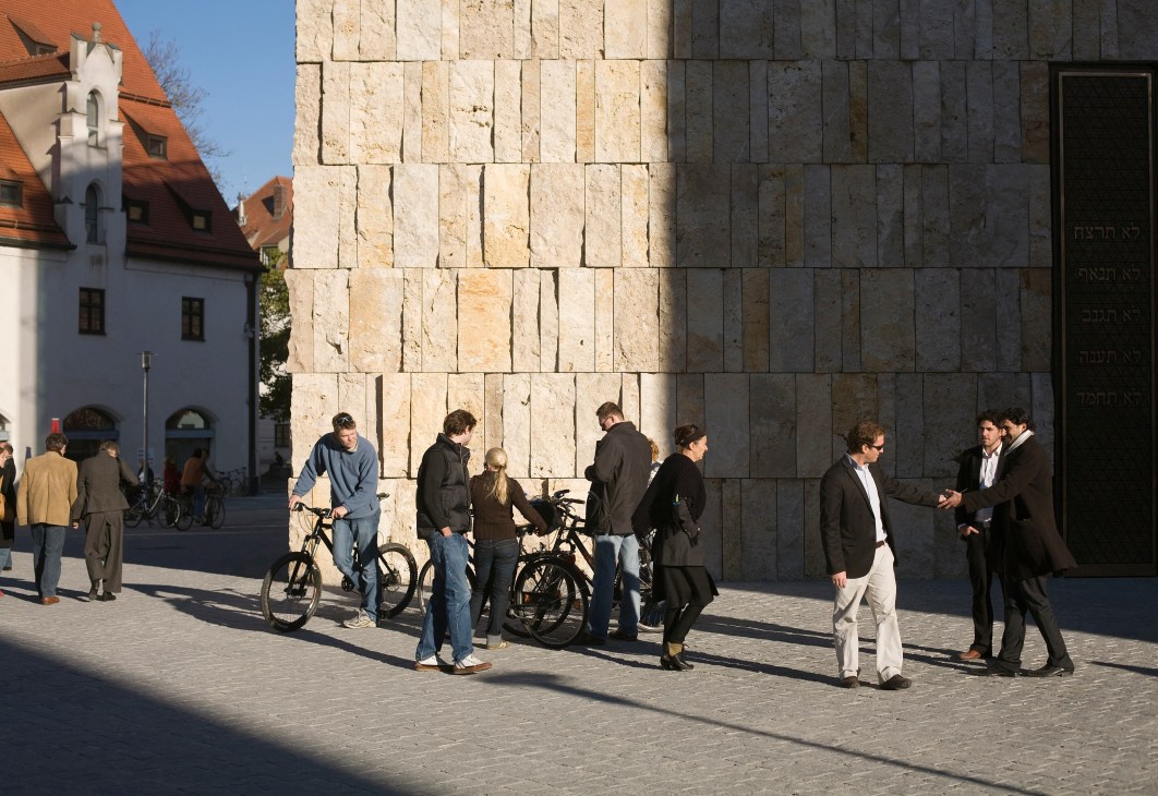 People with bicycles gather in a sunny square beside a large stone building with Hebrew text and a white chapel with red roof visible.