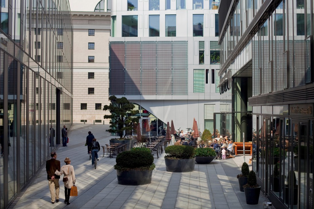 Modern courtyard with pedestrians, cyclists, and outdoor seating surrounded by contemporary glass and stone buildings.