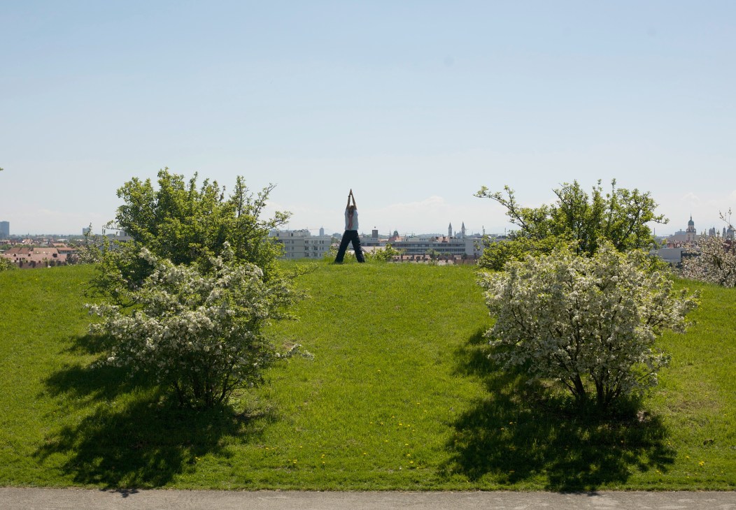 Person in yoga pose on green hillside with blooming trees overlooking Munich cityscape.