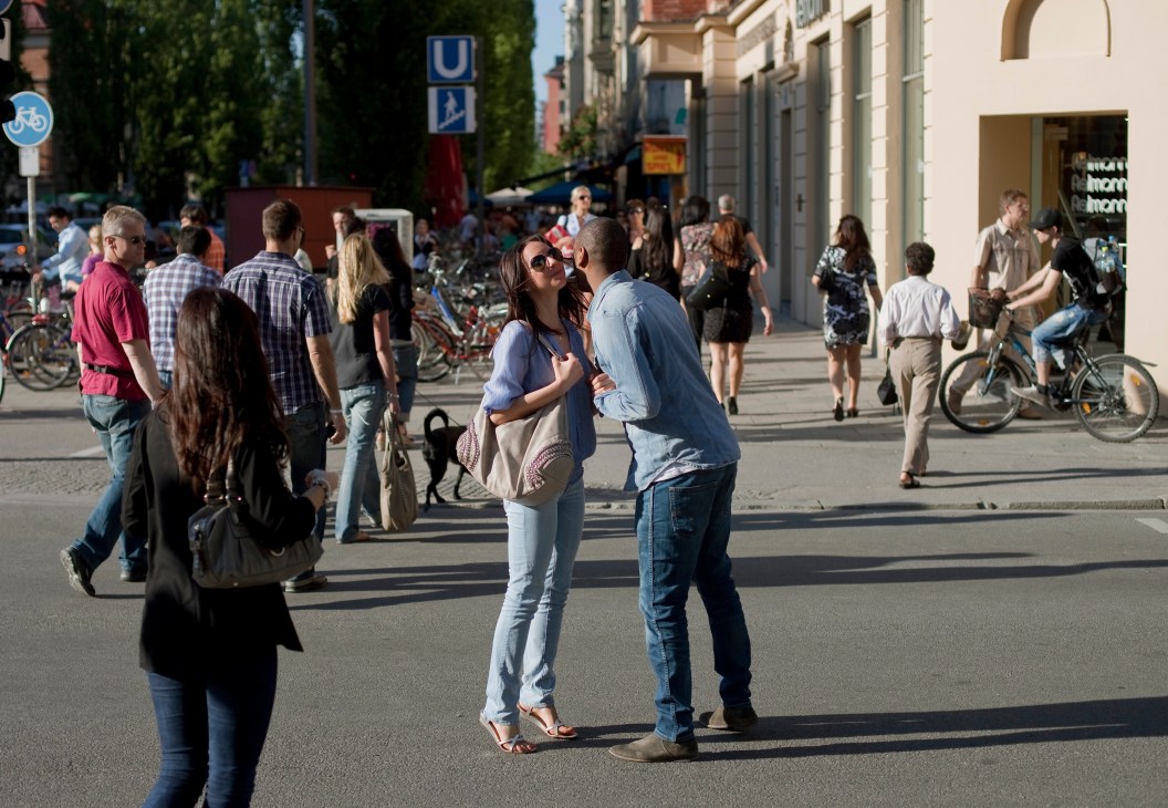 Pedestrians and cyclists on a sunny Munich street with shops and buildings lining the sidewalk.