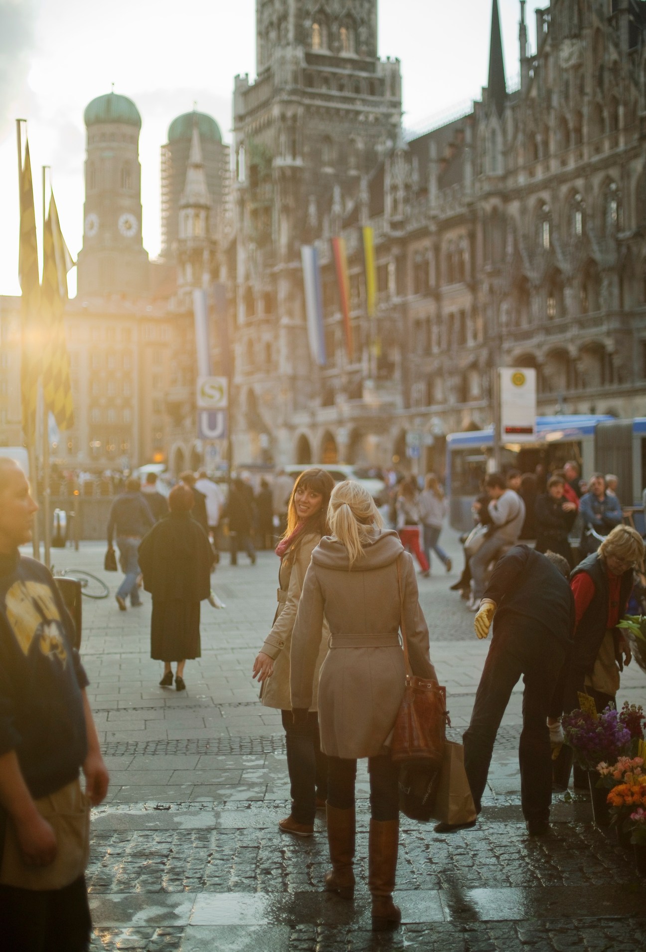 Woman with blonde hair in beige coat stands in Marienplatz square with Gothic city hall and twin church towers in background during golden hour.